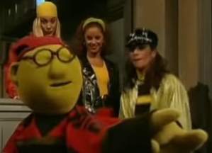 Foreground: Bunsen Honeydew on a red and black shirt and backwards red hat. Background: the three "Fly Girls" backup dancers.